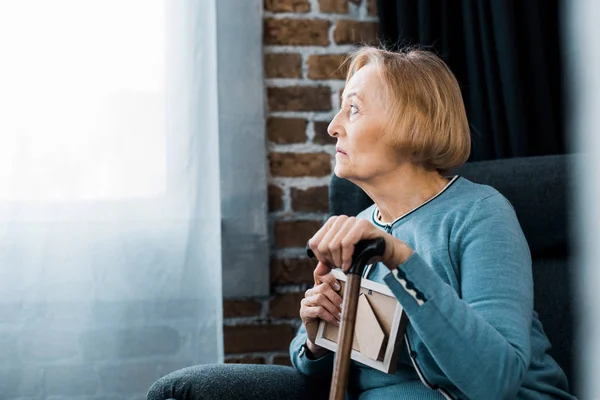 Upset senior woman sitting with walking stick and holding picture frame at home — Stock Photo