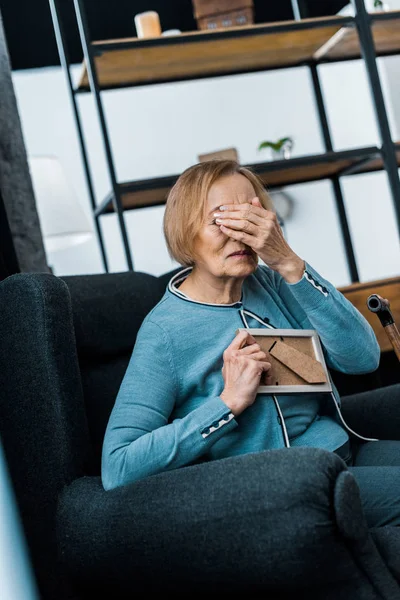 Grieving senior woman sitting in armchair, covering face with hand and crying while holding picture frame — Stock Photo