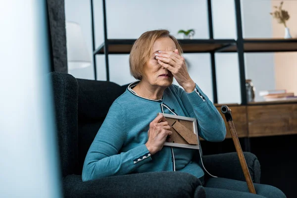 Trauernde Seniorin sitzt im Sessel, bedeckt Gesicht mit der Hand und weint, während sie den Bilderrahmen hält — Stockfoto
