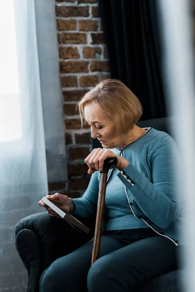 Upset senior woman sitting with walking stick and looking at picture frame — Stock Photo