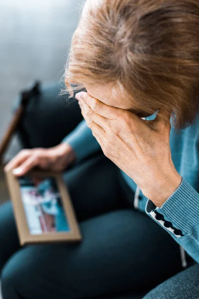 Mujer mayor cubriendo la cara con la mano y llorando mientras mira el marco de la foto - foto de stock