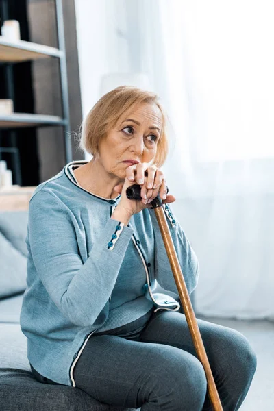 Sad senior woman sitting on couch and holding walking stick at home with copy space — Stock Photo
