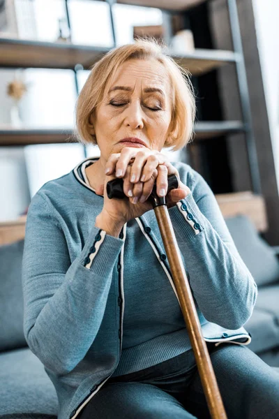 Sad senior woman with eyes closed leaning on walking stick at home — Stock Photo