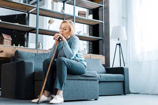 Traurige Seniorin sitzt auf Couch und lehnt an Gehstock im Wohnzimmer — Stockfoto