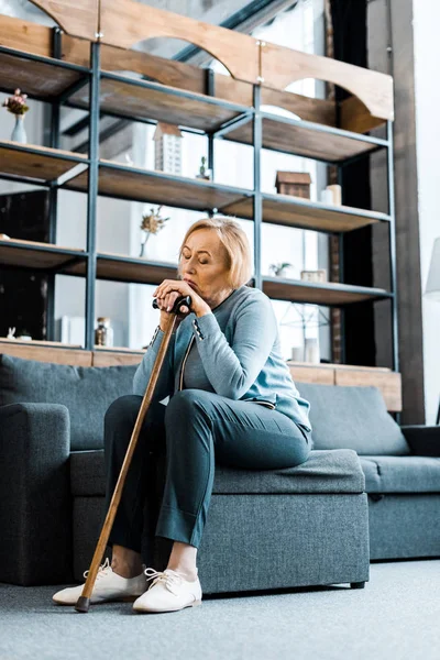 Sad senior woman sitting on couch and leaning on walking stick in living room — Stock Photo