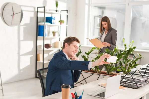 Enfoque selectivo de empresario feliz tomando selfie con compañera de trabajo en segundo plano - foto de stock