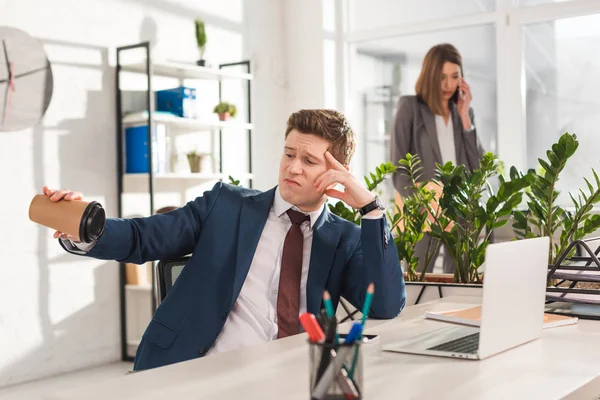 Selective focus of upset businessman looking at empty paper cup near female coworker on background — Stock Photo