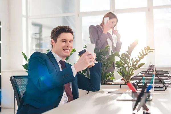 Foco seletivo de empresário alegre segurando smartphone e assistir vídeo perto de colega de trabalho feminino em segundo plano — Fotografia de Stock