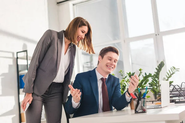 Cheerful businessman holding smartphone and watching video with female coworker — Stock Photo