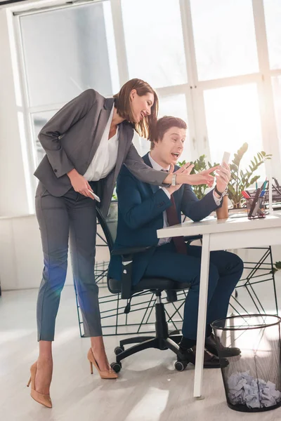 Attractive businesswoman gesturing near excited coworker holding smartphone and watching video — Stock Photo