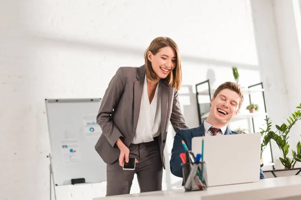 Приваблива бізнес-леді, що стоїть зі смартфоном біля щасливого колеги і сміється під час перегляду відео на ноутбуці — стокове фото
