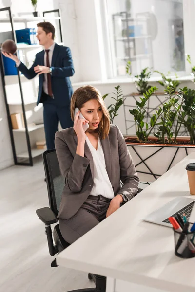 Messa a fuoco selettiva di attraente donna d'affari che parla su smartphone con collega sullo sfondo — Foto stock