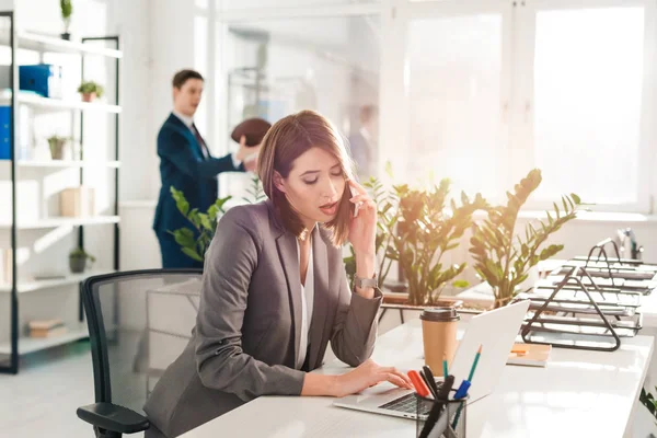 Selektive Fokussierung der Geschäftsfrau auf Smartphone in der Nähe von Laptop und Pappbecher mit Mitarbeiterin im Hintergrund — Stockfoto
