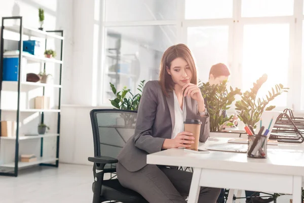 Enfoque selectivo de la empresaria pensativa sosteniendo taza de papel cerca de la computadora portátil con su compañero de trabajo en el fondo - foto de stock