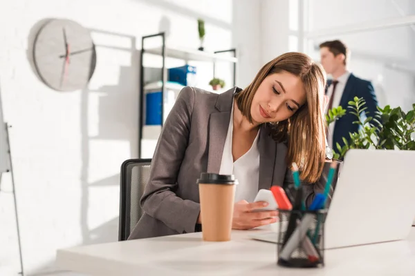 Selective focus of pensive businesswoman holding smartphone near paper cup and laptop with coworker on background — Stock Photo