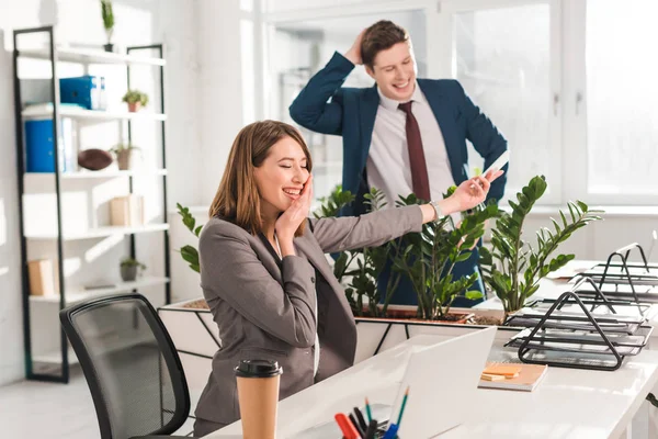 Femme d'affaires heureuse toucher le visage tout en tenant smartphone et rire avec un collègue au bureau — Photo de stock