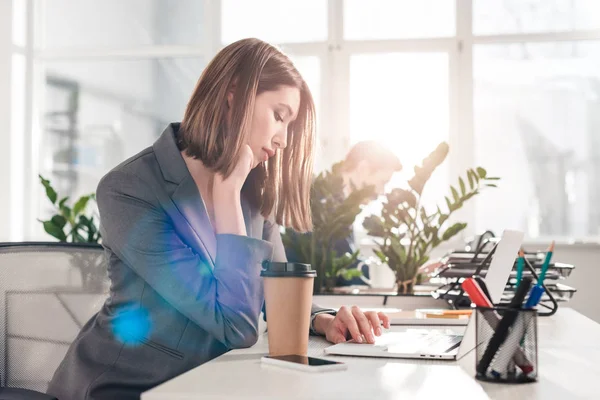 Enfoque selectivo de la mujer de negocios pensativa utilizando el ordenador portátil cerca de la taza de papel con su compañero de trabajo en el fondo - foto de stock