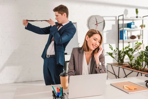 Atractiva mujer de negocios sonriendo mientras habla en el teléfono inteligente cerca de compañero de trabajo en la oficina, concepto de dilación - foto de stock