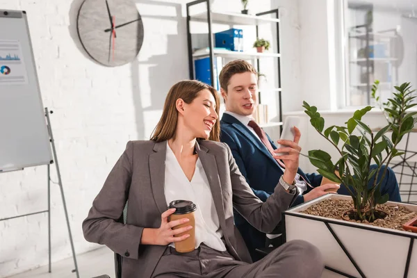 Attractive businesswoman smiling while holding paper cup and smartphone near coworker in office — Stock Photo