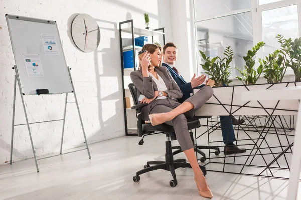 Attractive businesswoman smiling while holding paper cup and talking with coworker in office,  procrastination concept — Stock Photo