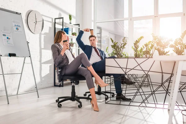 Alegre mujer de negocios sosteniendo taza de papel y hablando con compañero de trabajo sosteniendo en la cabeza taza desechable en la oficina, concepto de dilación - foto de stock