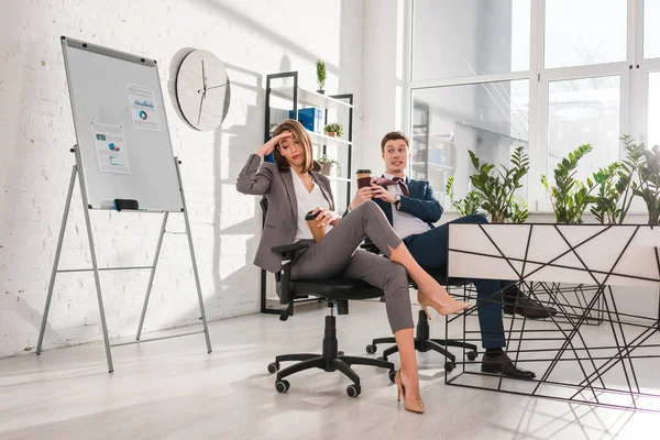 Mujer de negocios cansado sosteniendo taza de papel y sentado cerca de compañero de trabajo en la oficina - foto de stock