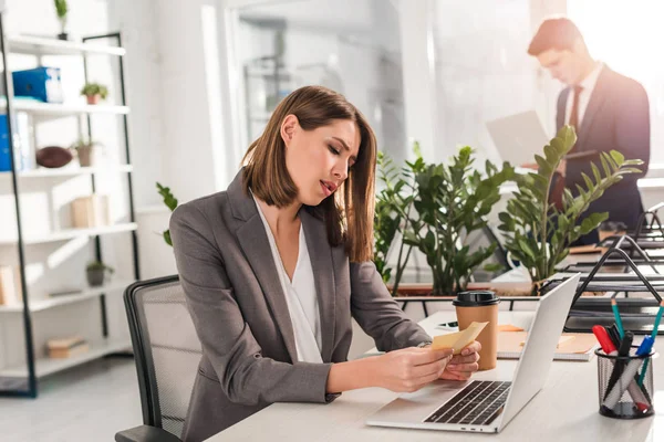 Selective focus of attractive businesswoman looking at sticky notes near laptop with coworker on background — Stock Photo