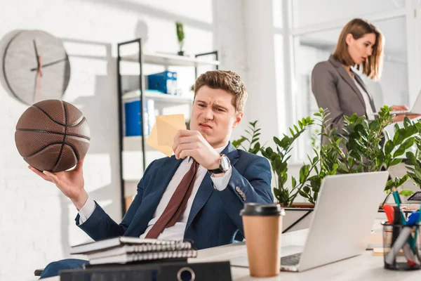 Selektiver Fokus des unzufriedenen Geschäftsmannes, der auf klebrige Zettel blickt, während er Basketball im Amt hält, mit Mitarbeiter auf Hintergrund, Zauderungskonzept — Stockfoto