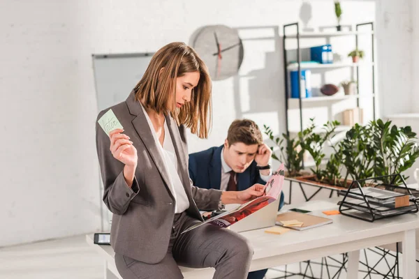 Selective focus of woman holding sticky note with later lettering and reading magazine with coworker sitting at desk on background, procrastination concept — Stock Photo