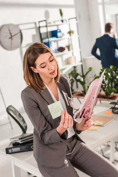 Selective focus of businesswoman holding sticky note with later lettering and reading magazine with coworker on background, procrastination concept — Stock Photo