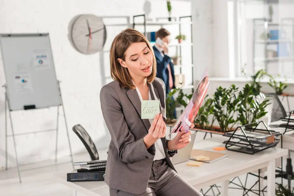 Enfoque selectivo de la mujer de negocios sosteniendo nota adhesiva con letras posteriores y revista en las manos con su compañero de trabajo en el fondo, concepto de dilación - foto de stock