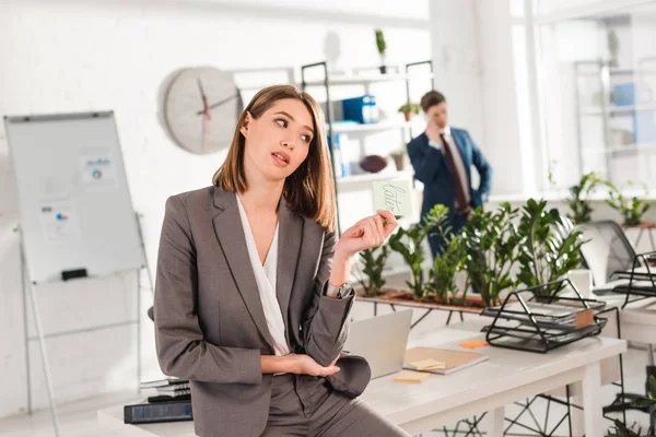 Messa a fuoco selettiva della donna d'affari che tiene nota appiccicosa con scritte successive in mano con il collega sullo sfondo, concetto di procrastinazione — Foto stock