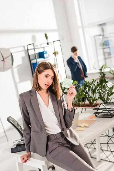 Enfoque selectivo de la mujer de negocios sosteniendo nota adhesiva con letras posteriores con compañero de trabajo en el fondo, concepto de dilación - foto de stock