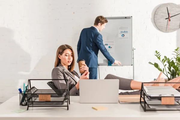 Selektiver Fokus der schönen Geschäftsfrau, die Pappbecher in der Hand hält und neben dem Laptop ruht, mit einem Mitarbeiter im Hintergrund — Stockfoto