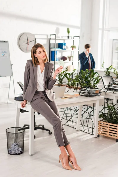 Enfoque selectivo de la mujer sosteniendo nota adhesiva con letras posteriores, mientras que de pie cerca del escritorio con su compañero de trabajo en el fondo, el concepto de dilación - foto de stock