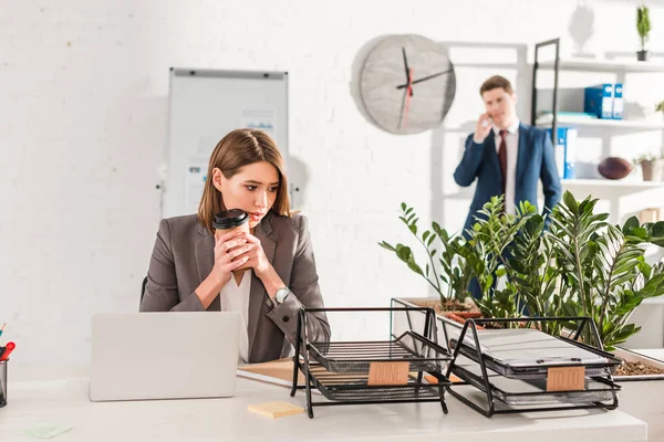 Selective focus of beautiful businesswoman holding paper cup and looking at document trays with lettering near coworker — Stock Photo