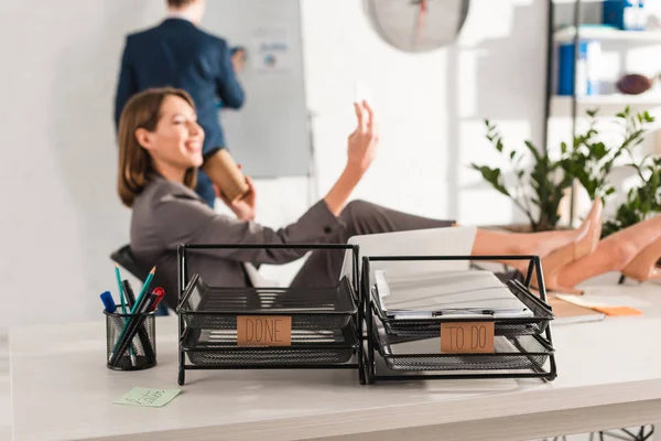 Selective focus of document trays with lettering near businesswoman taking selfie with paper cup, procrastination concept — Stock Photo