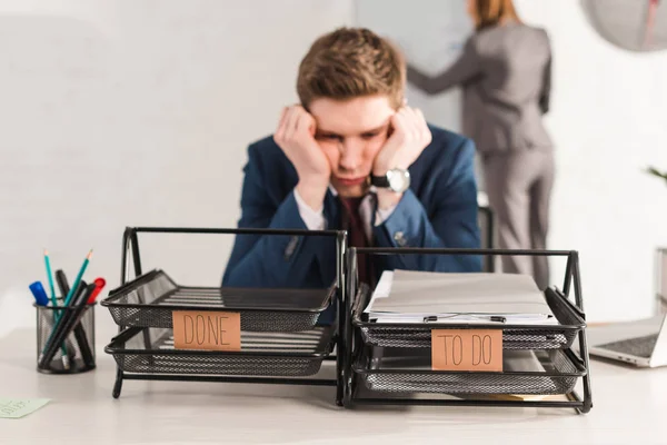 Selective focus of document trays with lettering near tired man and female coworker on background, procrastination concept — Stock Photo
