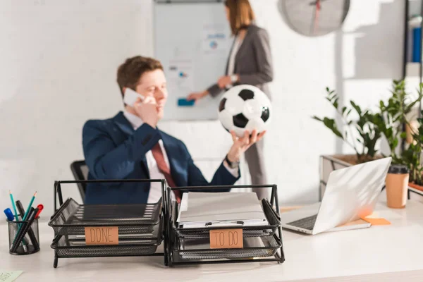 Selective focus of document trays with lettering near businessman talking on smartphone and holding football near female coworker on background, procrastination concept — Stock Photo