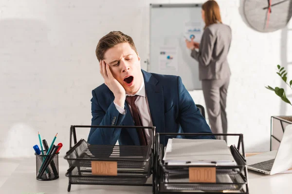 Selective focus of tired man yawning near document trays with lettering with female coworker on background, procrastination concept — Stock Photo