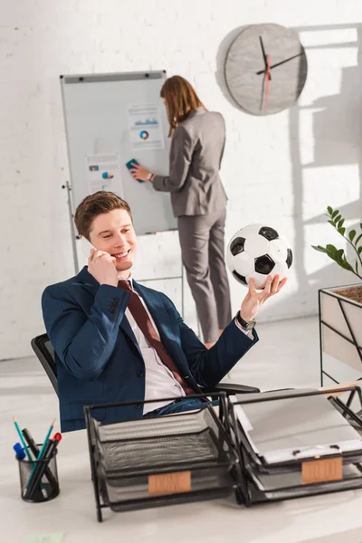 Selektiver Fokus des Geschäftsmannes, der auf dem Smartphone spricht und Fußball in der Nähe von Aktenschubladen mit Schriftzug hält und weibliche Mitarbeiterin im Hintergrund, Zauderkonzept — Stockfoto