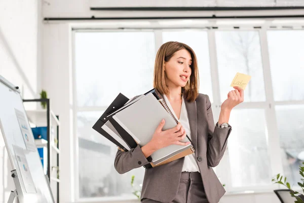 Atractiva mujer de negocios sosteniendo carpetas y mirando nota adhesiva con letras posteriores en la oficina - foto de stock