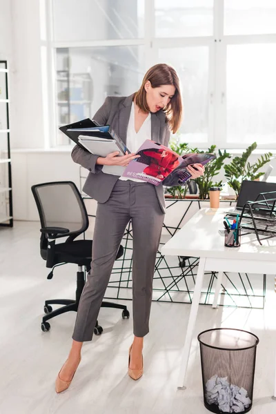 Séduisante femme d'affaires tenant des dossiers et lisant le magazine tout en restant au bureau — Photo de stock
