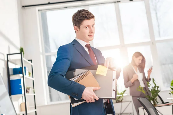 Guapo hombre de negocios sosteniendo carpetas y nota adhesiva con letras posteriores en la oficina con su compañera de trabajo en segundo plano - foto de stock