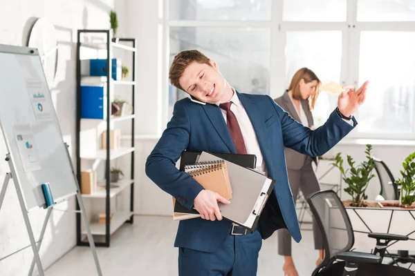 Handsome businessman holding folders and talking on smartphone in office with female coworker on background — Stock Photo