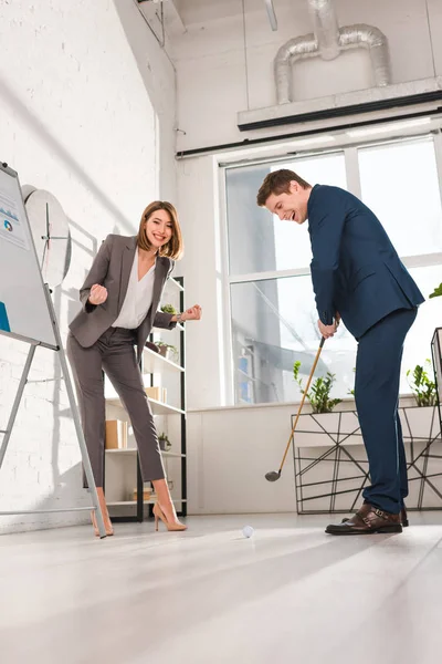 Cheerful woman gesturing while standing near handsome businessman playing mini golf in office — Stock Photo