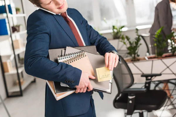 Cropped view of businessman holding folders and sticky note with later lettering and talking on smartphone in office — Stock Photo