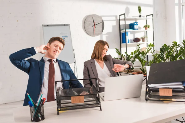 Selective focus of document trays with lettering near businesswoman looking at watch near tired man, procrastination concept — Stock Photo