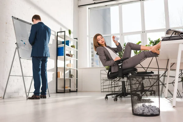 Selektiver Fokus der attraktiven Geschäftsfrau, die auf dem Stuhl sitzt und Papier in den Papierkorb neben dem Mitarbeiter wirft, Aufschiebekonzept — Stockfoto