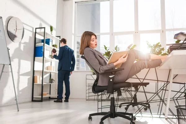 Selektiver Fokus der attraktiven Geschäftsfrau, die sich auf dem Stuhl ausruht und in der Nähe ihres Mitarbeiters auf das Notizbuch blickt — Stockfoto